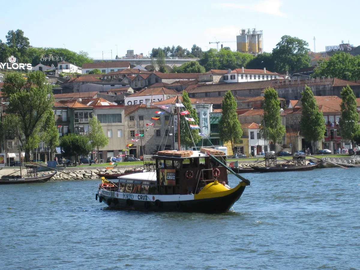 Port wine lodges with rooftop signs on the Gaia riverfront behind a rabelo boat, a port cellar day trip from Porto